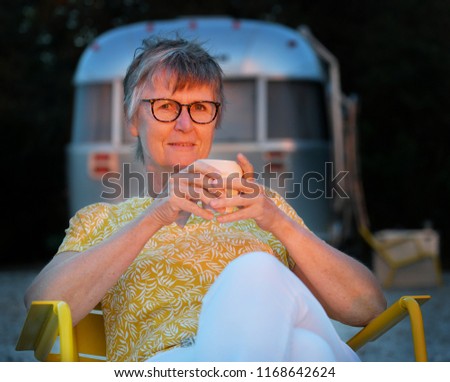 An older woman with glasses holding a cup of coffee is sitting in a yellow chair with her face in the last sunlight in front of an airstream Travel Trailer