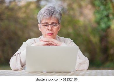 Older Woman With Gray Hair In A Park With A Gray Computer Sits On A Chair In The Autumn Outdoor Ambience