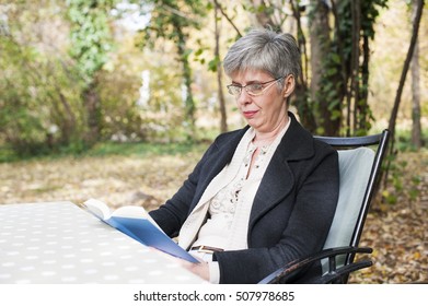 Older Woman With Gray Hair Sitting In The Park Reading A Book