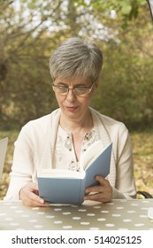 Older Woman With Gray Hair Sitting In The Park Reading A Book