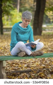 Older Woman With Gray Hair Sitting On Bench In The Park Reading A Book