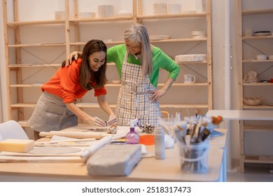 Older woman guiding a young woman through pottery techniques in a friendly, creative studio. - Powered by Shutterstock