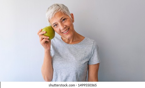 Older Woman With Healthy Food Indoors. Smiling Woman Eating Green Apple. Portrait Of A Beautiful Elderly Woman Holding An Apple, Smiling, Isolated On Gray Background.