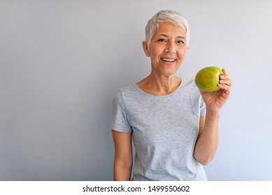 Older Woman With Healthy Food Indoors. Smiling Woman Eating Green Apple. Portrait Of A Beautiful Elderly Woman Holding An Apple, Smiling, Isolated On Gray Background.