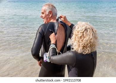 Older Woman Helping Her Husband To Put On His Wetsuit On The Beach For A Swimming Trip