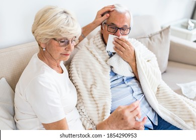 An Older Woman Helps A Ill Husband While They Are Sitting On The Couch In The Living Room. Senior Man With Flu And Running Nose.