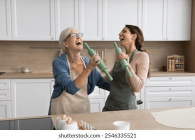 Older woman her young adult daughter hold rolling-pins and singing together in modern kitchen, having fun, fooling around, while cooking homemade pastries on weekend leisure or family holiday at home - Powered by Shutterstock