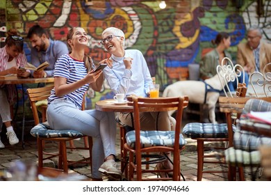 An Older Woman And Her Young Female Friend Laughing About A Smartphone Content They Watching While Have A Drink In A Cheerful Atmosphere In The Bar. Leisure, Bar, Friendship, Outdoor