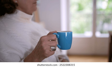 Older woman holds a blue mug, enjoying a quiet moment, with natural light streaming through the window, emphasizing comfort, relaxation, and the simple pleasures of life - Powered by Shutterstock