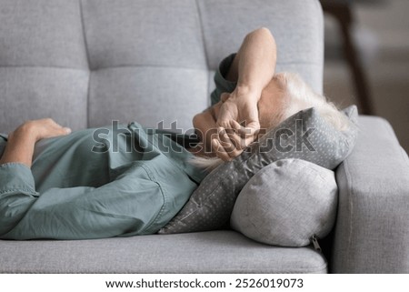 Older woman lying on sofa cushion, feeling fatigued, unmotivated, taking rest at home. Peaceful aged female having midday break, daytime nap indoors, relieving stress and mental or physical exhaustion