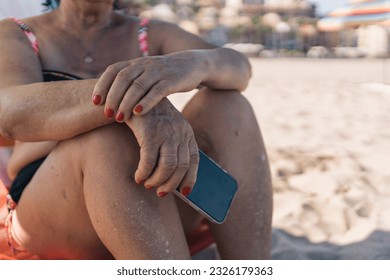 Older woman on the beach holding her mobile phone. Elderly lady on the beach sitting and relaxing, dropping her hands to hold her mobile phone. - Powered by Shutterstock