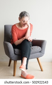 Older Woman In Orange Top Seated In Armchair With Hand Over Eyes