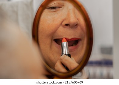 An Older Woman Paints Her Lips With Red Lipstick In Front Of A Mirror