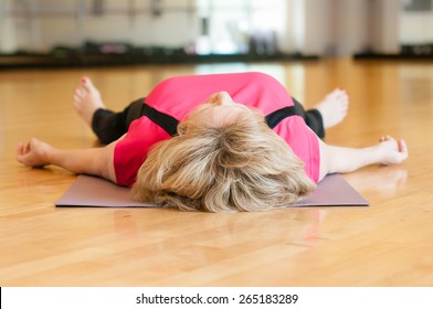 An Older Woman In A Pink Yoga Outfit Rests On The Floor Of The Gym Room In Savasana (corpse) Pose After Yoga Class,