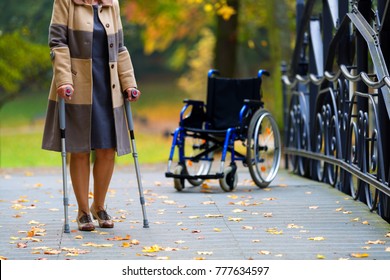older woman practicing walking on crutches in autumnal park with wheelchair standing in the background