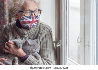 Older Woman In A Protective Mask With A British Flag And Cat Stays At Home Looking Out The Window. Lockdown In The UK. Coronavirus Mutated Covid-19. Strengthening Quarantine. Health Of The Elderly