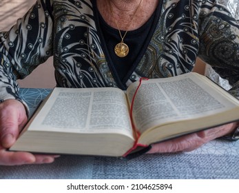 Older Woman Reading The Bible With A Gold Medallion Of The Virgin Mary Hanging Around Her Neck. Concept Of Christianity