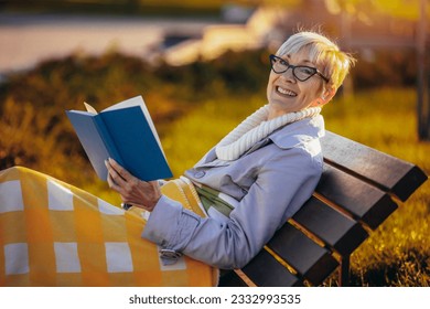 An older woman reading a book. She is sitting on the bench in the colorful park in the autumn - Powered by Shutterstock