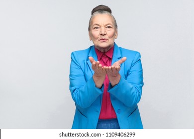 Older Woman Sending Air Kiss At Camera. Emotion And Feelings Handsome Expressive Grandmother With Light Blue Suit And Pink Shirt With Collected Bun Gray Hair. Studio Shot, Isolated On Gray Background