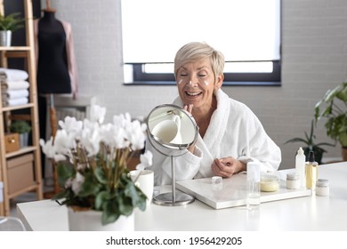 An Older Woman With Short Blonde Hair In A White Bathrobe Sits At The Table And Puts Cream On Her Face Inside The Living Room