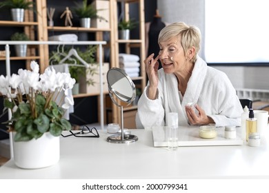 An Older Woman With Short Blonde Hair In A White Bathrobe Sits At The Table And Puts Cream On Her Face Inside The Living Room