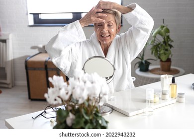 An older woman with short blonde hair in a white bathrobe sits at the table and puts cream on her face inside the living room