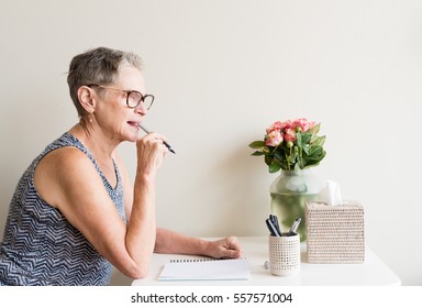 Older Woman With Short Grey Hair And Glasses Holding Pen Pensively At Writing Desk With Pink Roses (selective Focus)