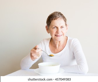 Older Woman With Short Hair And White Top Smiling And Eating Soup From A White Bowl