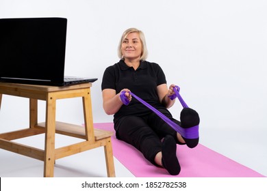 An Older Woman Sits On A Mat In Front Of A Computer And Does Exercises With An Elastic Band. Pandemic And Senior Online Concept. Photo On White Background