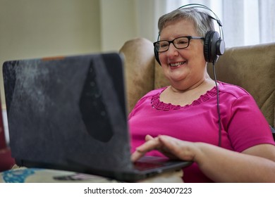 Older Woman Is Sitting In An Armchair Using Her Laptop. She Is Listening Music With A Black Headphones. She Wears Glasses And A Pink T-shirt. Behind Her, There Are Transparent Curtains.