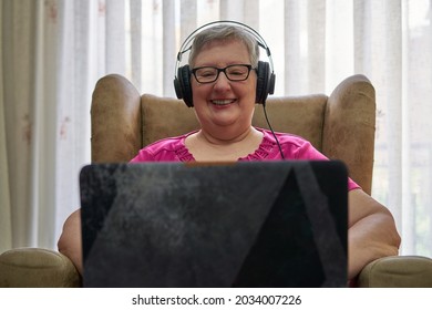 Older Woman Is Sitting In An Armchair Using Her Laptop. She Is Listening Music With A Black Headphones. She Wears Glasses And A Pink T-shirt. Behind Her, There Are Transparent Curtains.