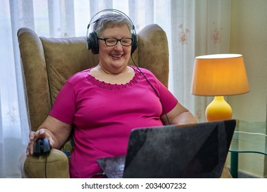 Older Woman Is Sitting In An Armchair Using Her Laptop. She Is Listening Music With A Black Headphones. She Is Holding The Mouse With Her Right Hand. She Wears Glasses And A Pink T-shirt.