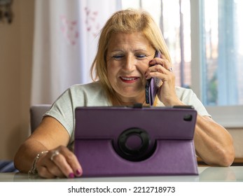 Older Woman Sitting At Home While Talking On Her Cell Phone With Someone Else And Is Entertaining Herself With Her Tablet.