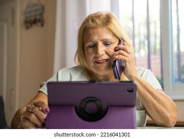 Older Woman Sitting At Home While Talking On Her Cell Phone With Someone Else And Is Entertaining Herself With Her Tablet.