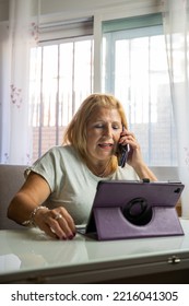 Older Woman Sitting At Home While Talking On Her Cell Phone With Someone Else And Is Entertaining Herself With Her Tablet.