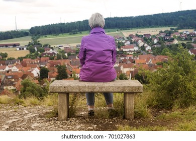 Older Woman Sitting On Bench And Looking Towards A Small Town