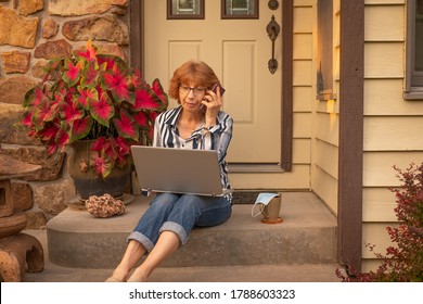 Older Woman Sitting On Front Porch Of Her House Working On Her Laptop Computer While Talking On The Phone At Sunset In Summer; A Mug And Face Protecting Mask Next To Her; Covid-19 In Midwest