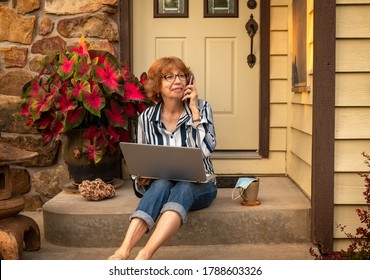 Older Woman Sitting On Front Porch Of Her House With A Laptop Computer Talking On The Phone And Smiling At Sunset In Summer; Beverage Cup And Face Protecting Mask Next To Her; Covid-19  In Midwest