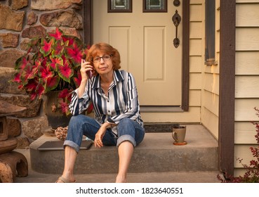 Older Woman Sitting On Front Porch Of Her House Talking On The Phone In Midwest, USA, In Summer;  Laptop Computer And Mug Next To Her