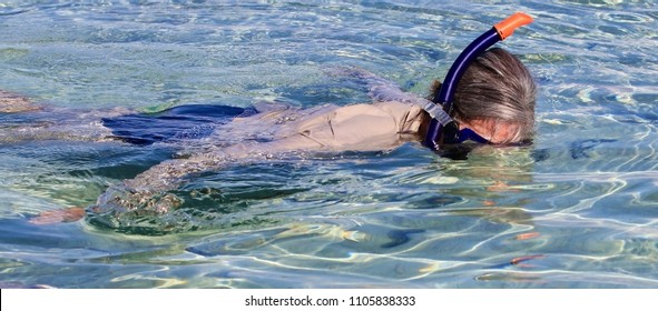 Older Woman Snorkeling Floating On The Surface Of The Clear, Turquoise  Ocean Water Wearing A Sunshirt.