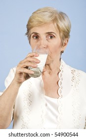 Older Woman Standing In Front Of Image,looking You And Drinking A Glass With Milk