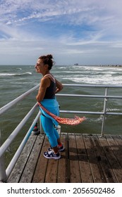 An Older Woman Standing On A Wooden Planked Boardwalk Looking Out At A Rough Ocean On A Windy Day With Her Scarf Flapping In The Wind