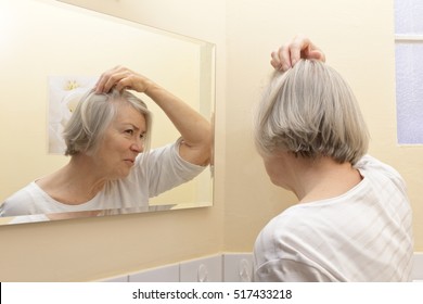 Older Woman With Thin Gray Hair And A Worried Look On Her Face  Examining Her Beginning Baldness  In The Mirror Of Her Yellow Bathroom 