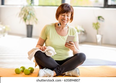 Older Woman Using Smartphone After Sports Training Sitting Indoors At Home Or Gym