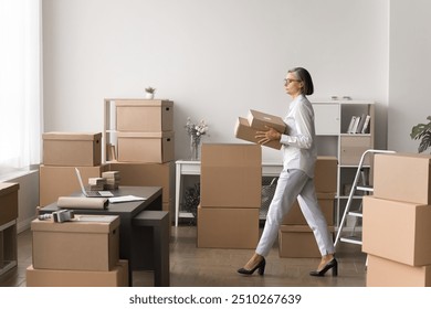 Older woman walking in warehouse room carrying small boxes, handling and moving parcels, overseeing and planning order dispatch, organizing items, busy in inventory management, logistics coordination - Powered by Shutterstock