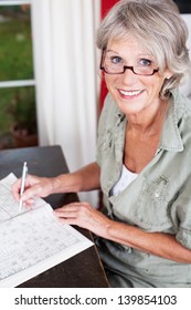 Older Woman Wearing Glasses Working On A Crossword Puzzle In A Puzzle Book Sittiing At A Small Wooden Table In Her House