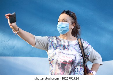 Older Woman Wearing A Medical Mask Taking A Selfie Near A Blue Wall.