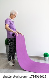 Older Woman With White Hair And Purple T-shirt Sportswear And Dark Tights Extending The Yoga Mat To Do Sport At Home
