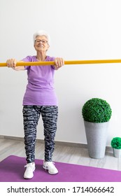 Older Woman With White Hair And Purple T-shirt Sportswear And Dark Tights Exercising At Home With A Pike