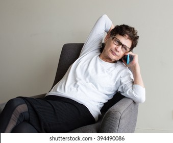 Older Woman With White Top And Glasses Using Smart Phone And Leaning Back In Chair